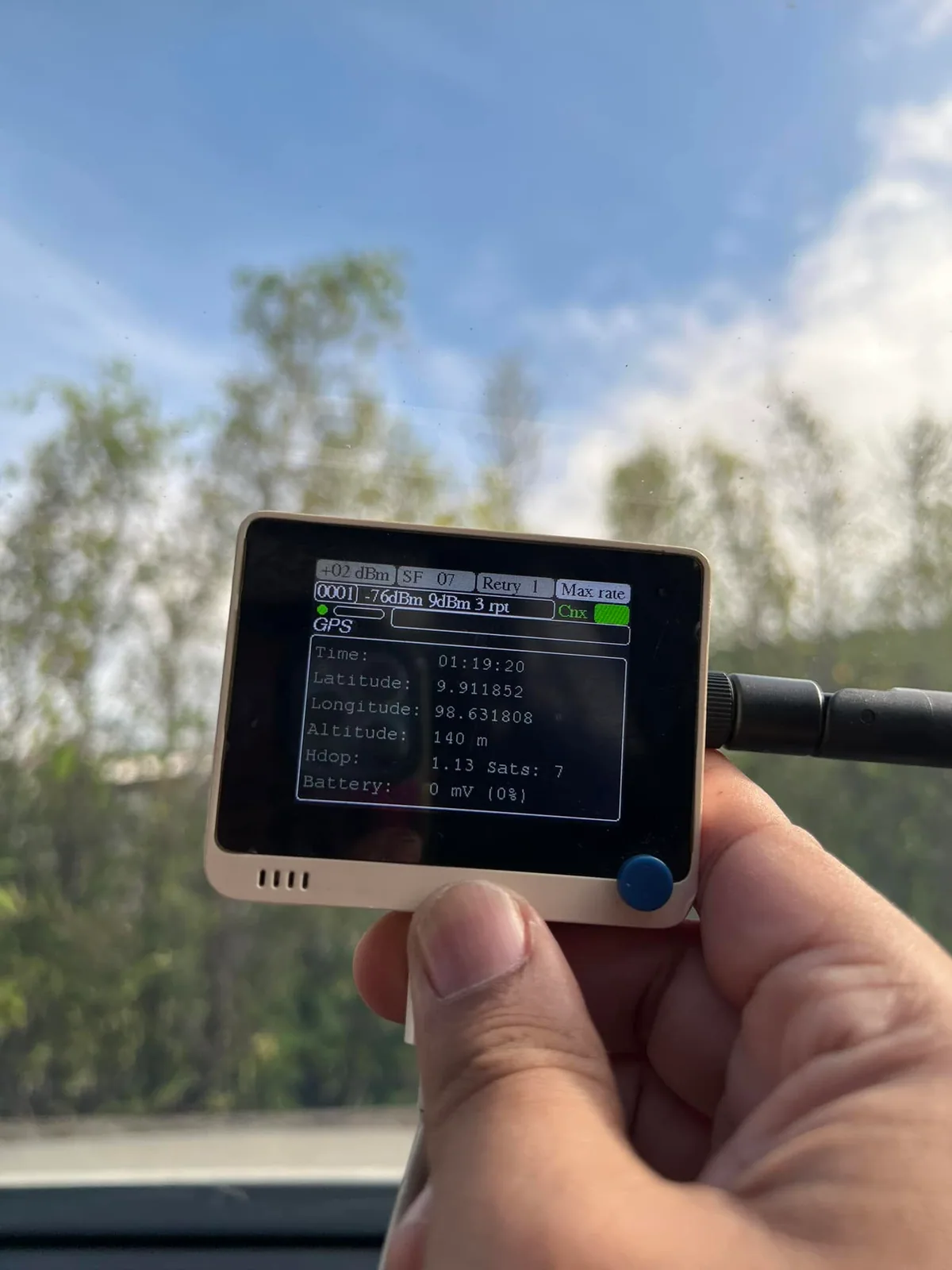 A hand holding a portable radio tester with signal and positioning information on its screen, outdoors.